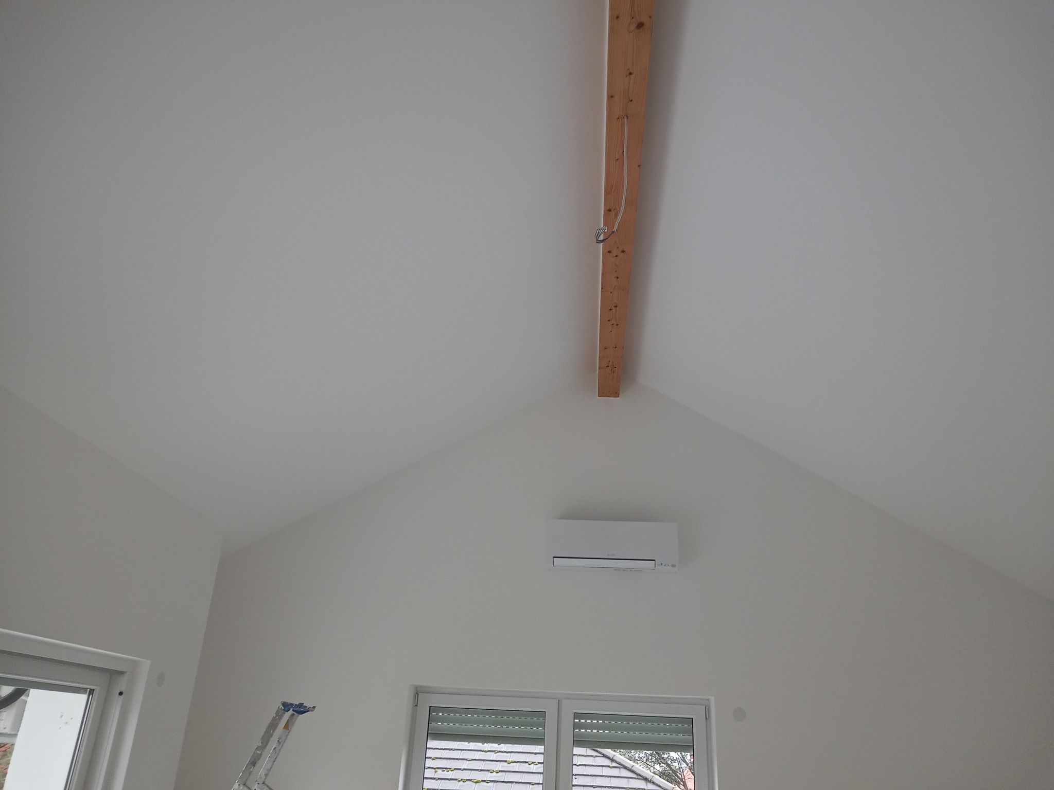 Poddasze z klimatyzatorem, pomalowane na biało, z widoczną drewnianą belką stropową i fragmentem drabiny malarskiej.