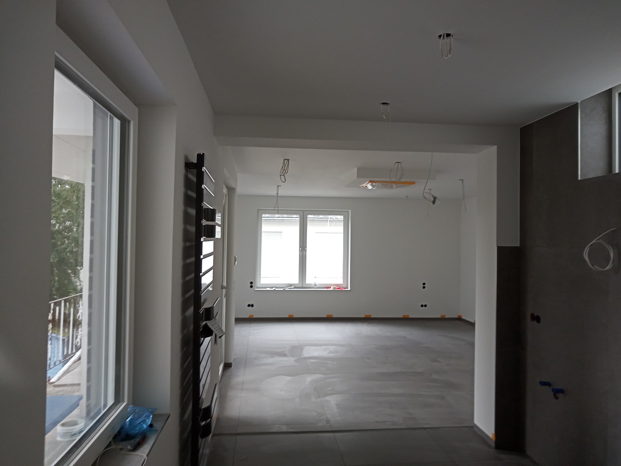 Wnętrze mieszkania w trakcie prac wykończeniowych: surowa podłoga, ściany przygotowane do malowania, widoczne kable i otwory na gniazdka, grzejnik naścienny, okno z widokiem na balkon.