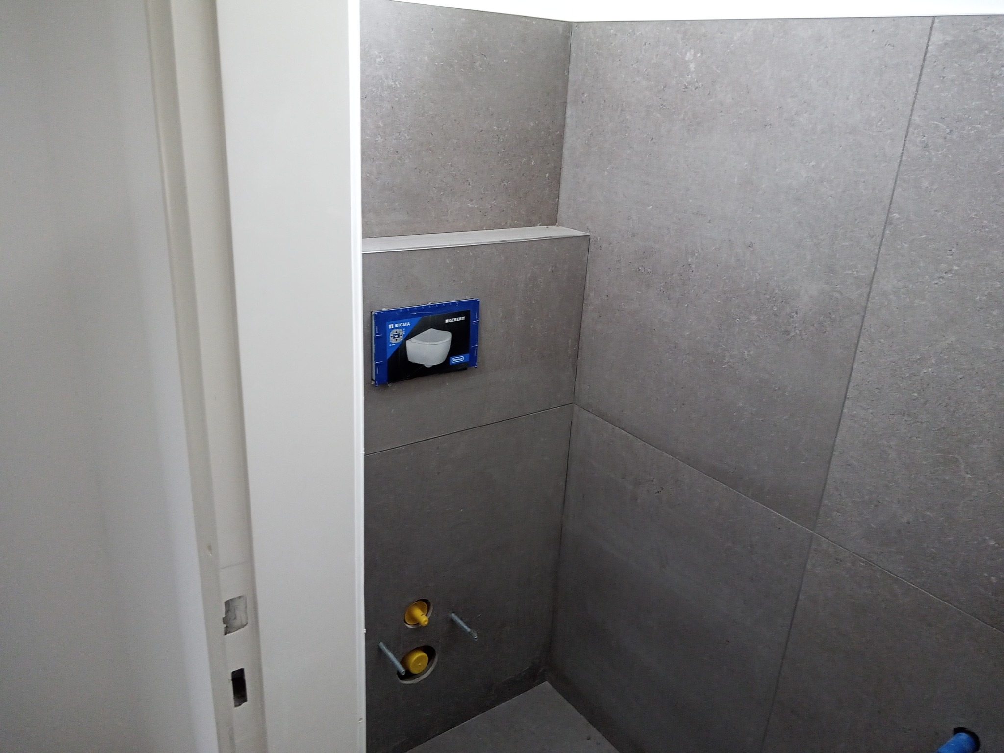 Wnęka w szarej łazience wyłożonej płytkami, z widocznym stelażem podtynkowym Geberit i instalacją hydrauliczną, z fragmentem białych drzwi po lewej stronie.