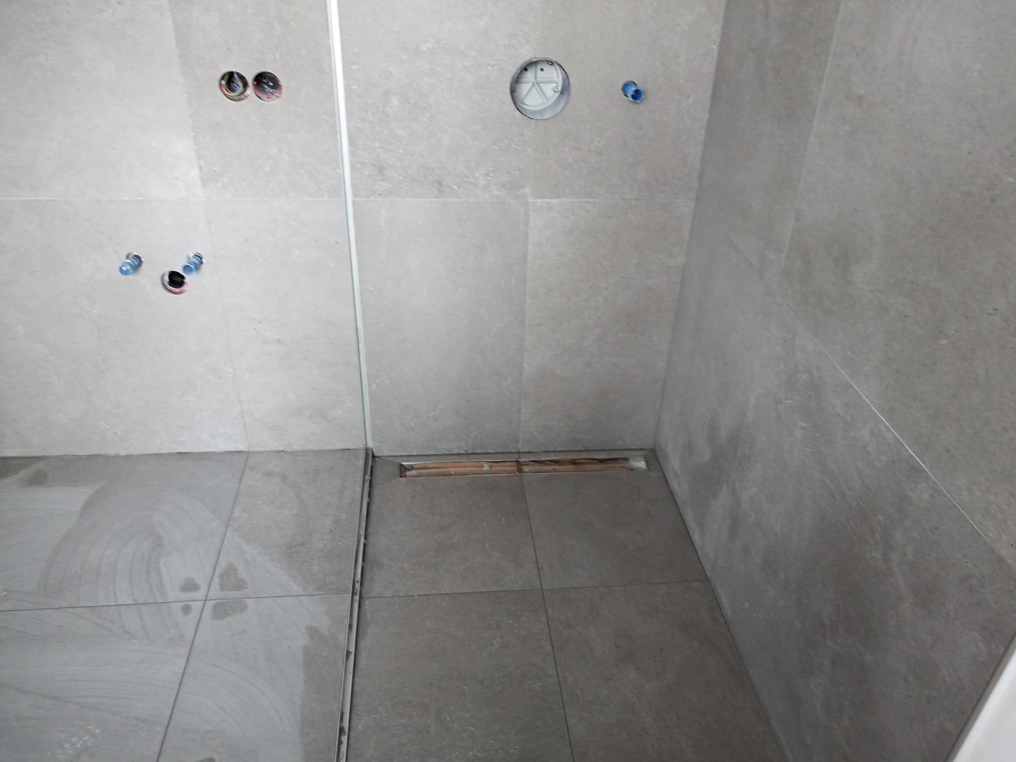 Szare płytki ścienne i podłogowe w surowej łazience w trakcie wykańczania, widoczne otwory na instalacje wodno-kanalizacyjne i odpływ liniowy.