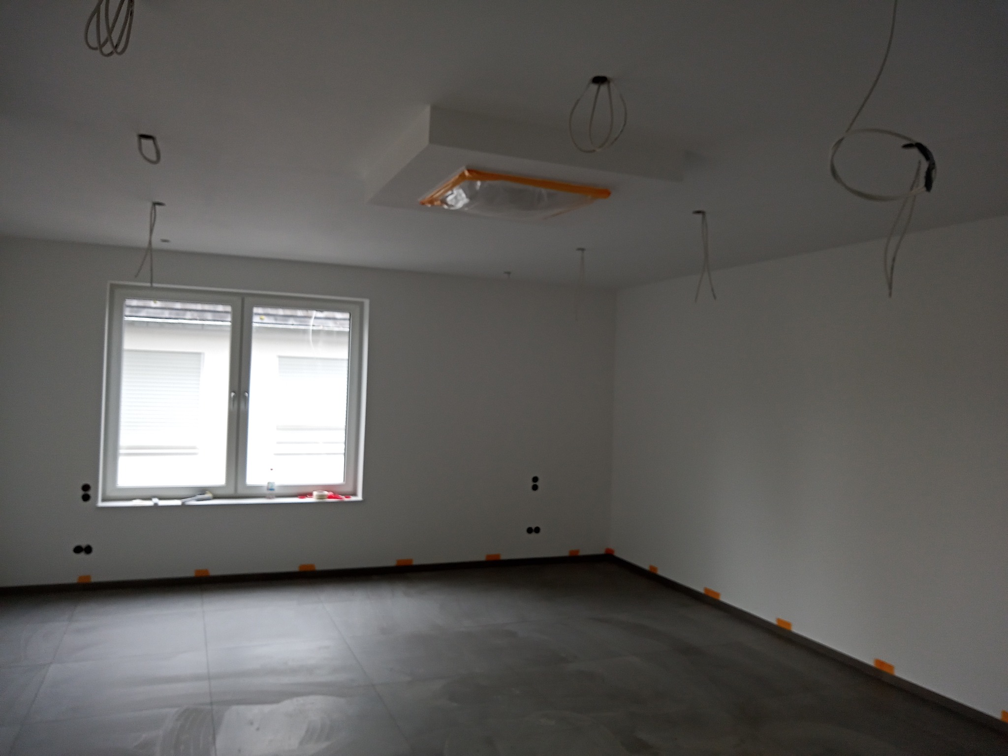 Pokój w trakcie remontu: szare płytki na podłodze, ściany pomalowane na biało, widoczne wystające kable, okno z żaluzjami, zabezpieczona lampa sufitowa.