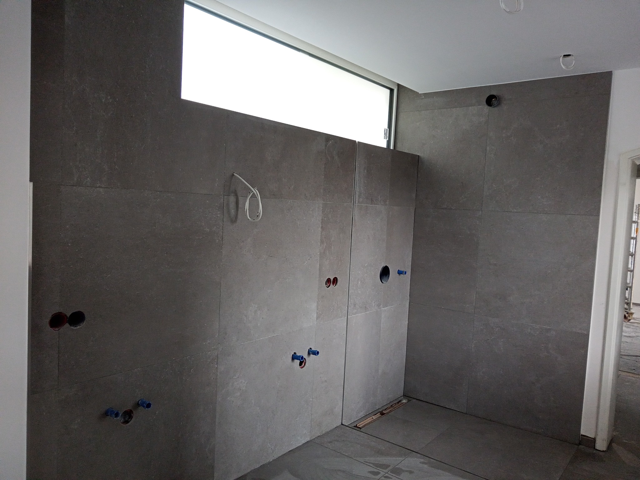 Wnętrze łazienki w trakcie wykańczania, szare płytki ścienne i podłogowe, widoczne otwory na instalacje wodne i elektryczne, okno pod sufitem i częściowo widoczna listwa odpływowa w podłodze.