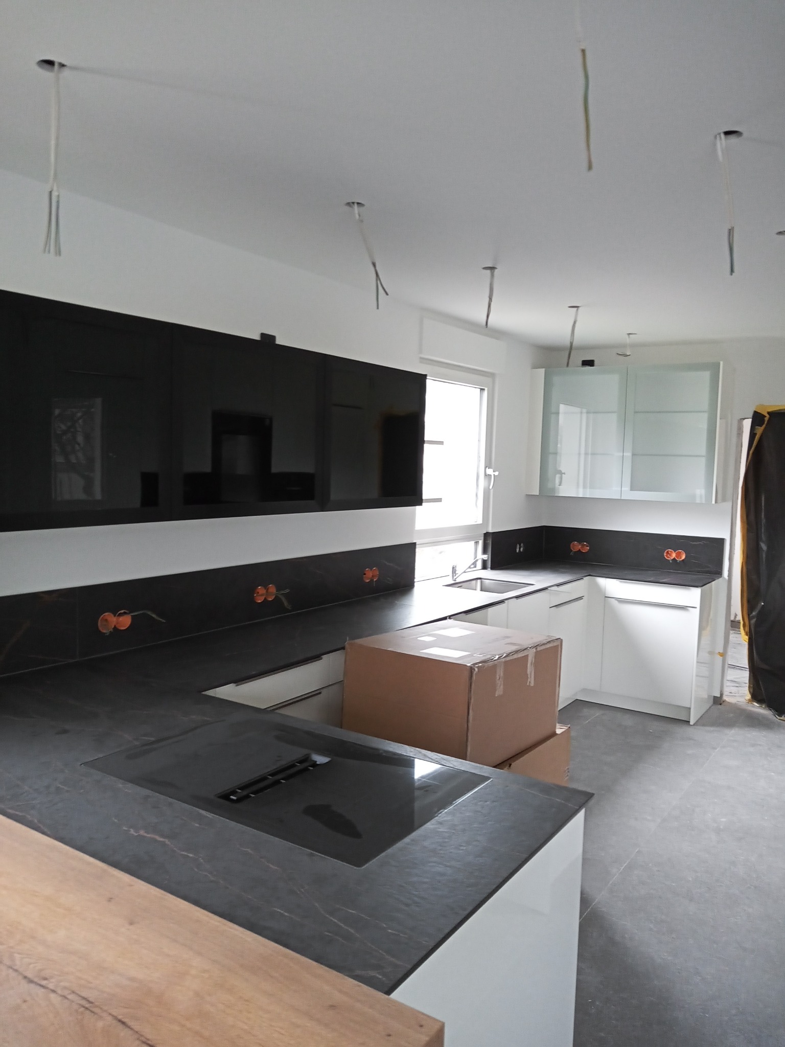 Wnętrze kuchni w trakcie montażu: czarne, błyszczące panele ścienne, szare blaty, białe szafki z przeszklonymi górnymi drzwiczkami, widoczne kable zwisające z sufitu, kartony na blacie.