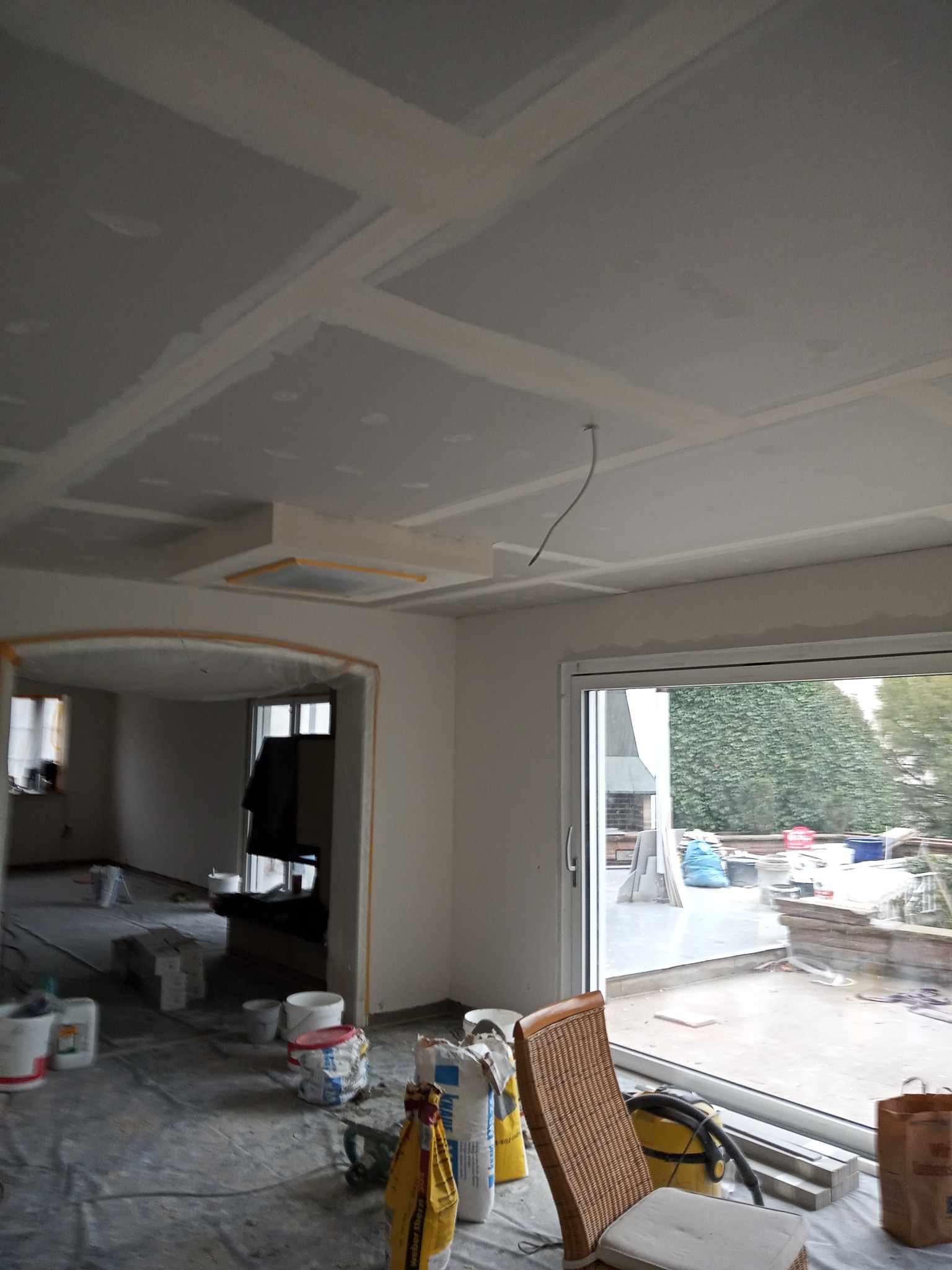 Wnętrze w trakcie remontu: sufit z płyt gipsowo-kartonowych ze szpachlowanymi łączeniami, folia ochronna na podłodze, worki z materiałami budowlanymi i krzesło wiklinowe w rogu pokoju.