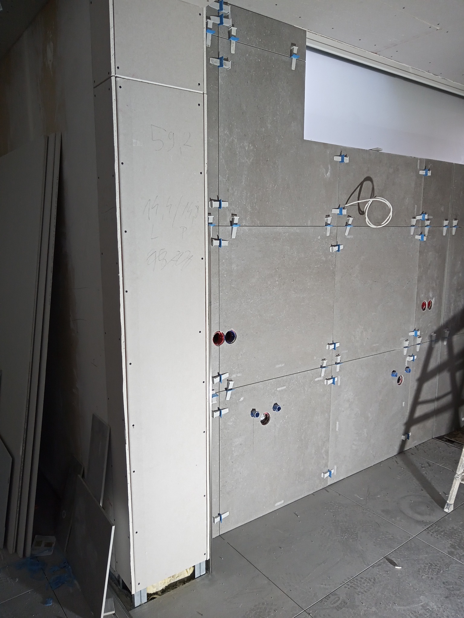 Szare płytki wielkoformatowe układane na ścianie z wykorzystaniem klipsów poziomujących, widoczne otwory pod instalacje i zapiski na płycie gipsowej.