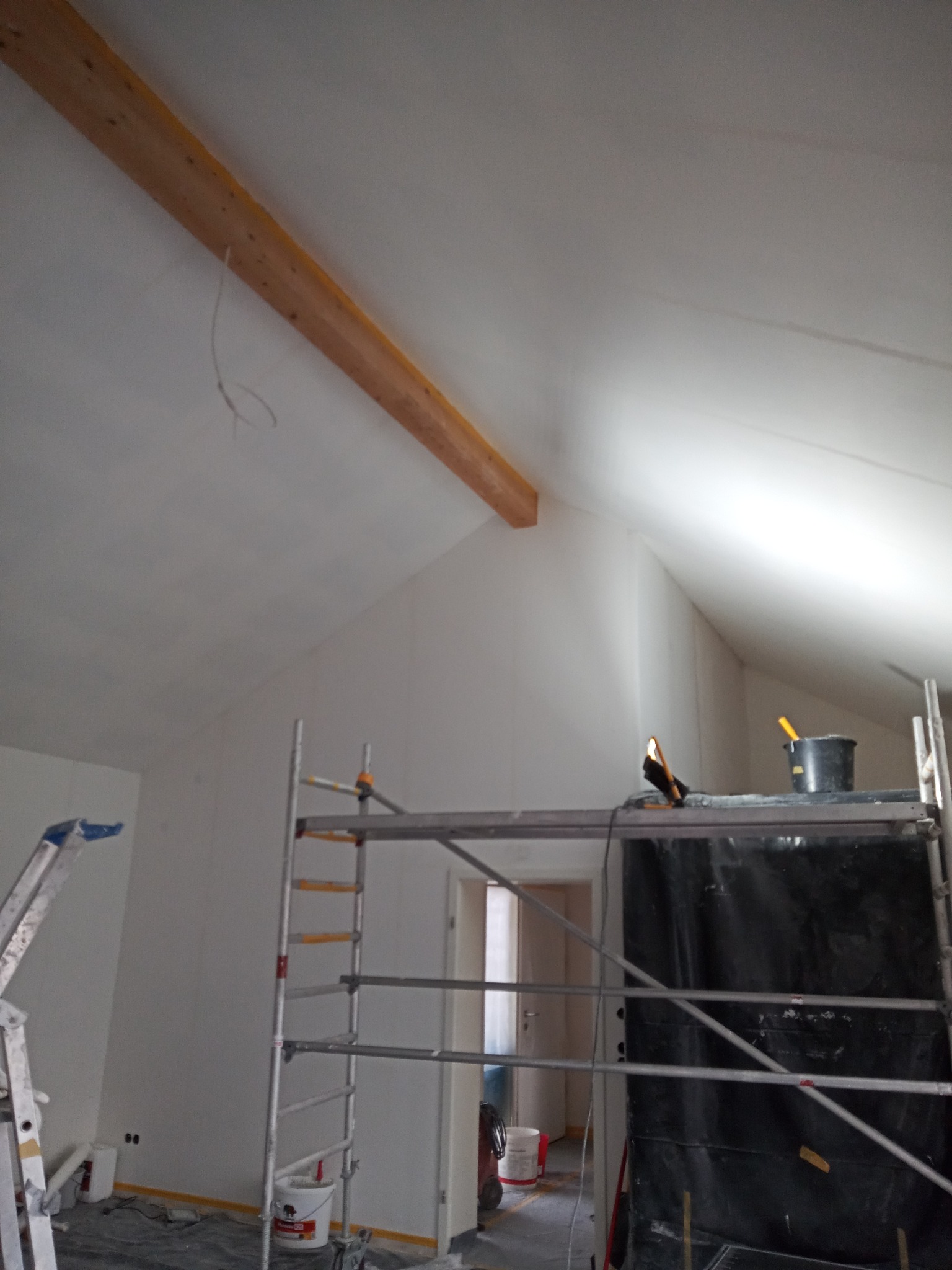 Pokój na poddaszu w trakcie malowania, widoczne rusztowanie, drabina i zabezpieczona folią ściana, sufit pomalowany na biało, widoczna drewniana belka stropowa.