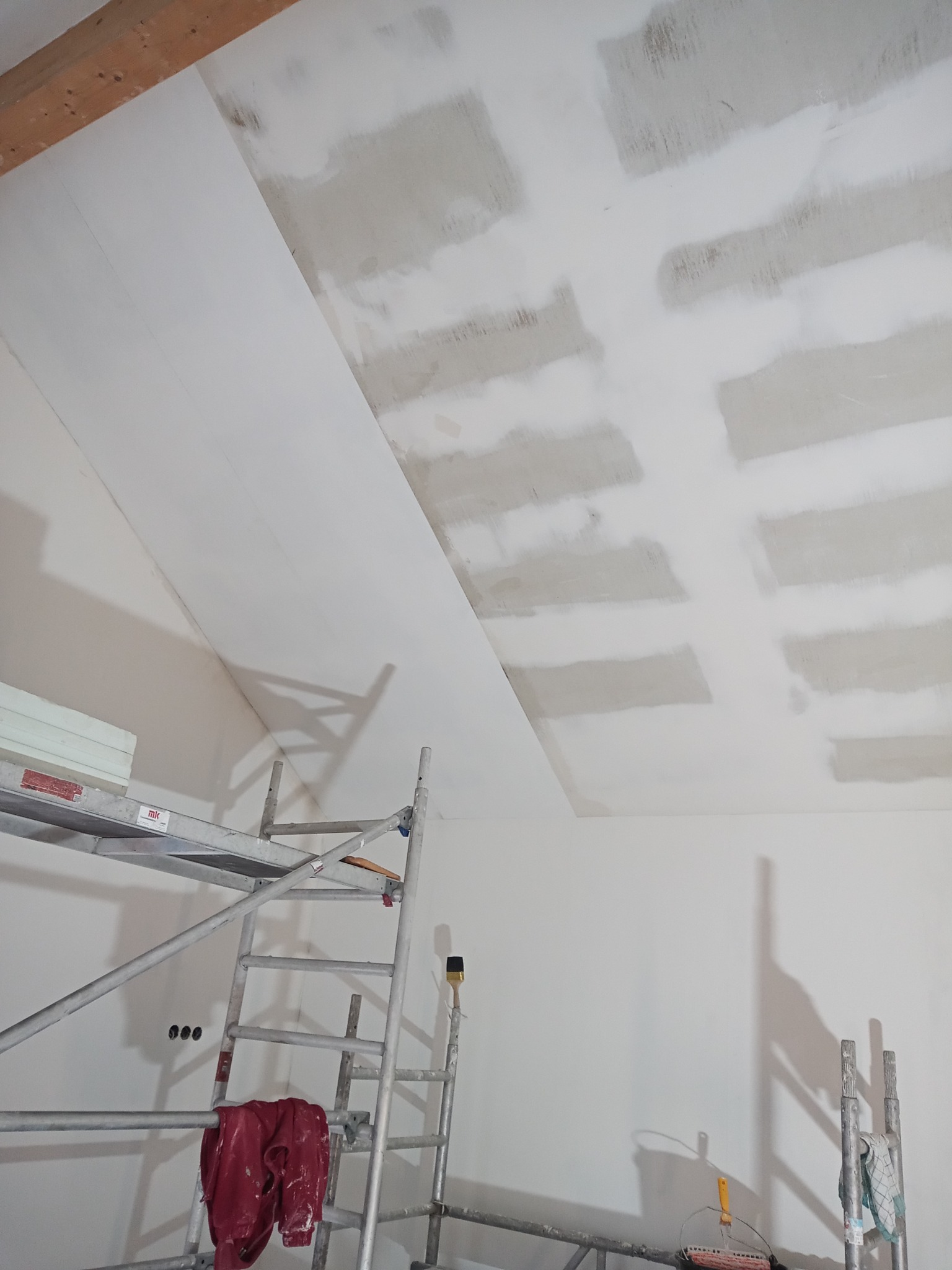 Sufit poddasza w trakcie szpachlowania łączeń płyt gipsowo-kartonowych, widoczne rusztowanie malarskie, ślady po szpachli i niezamalowane fragmenty sufitu.