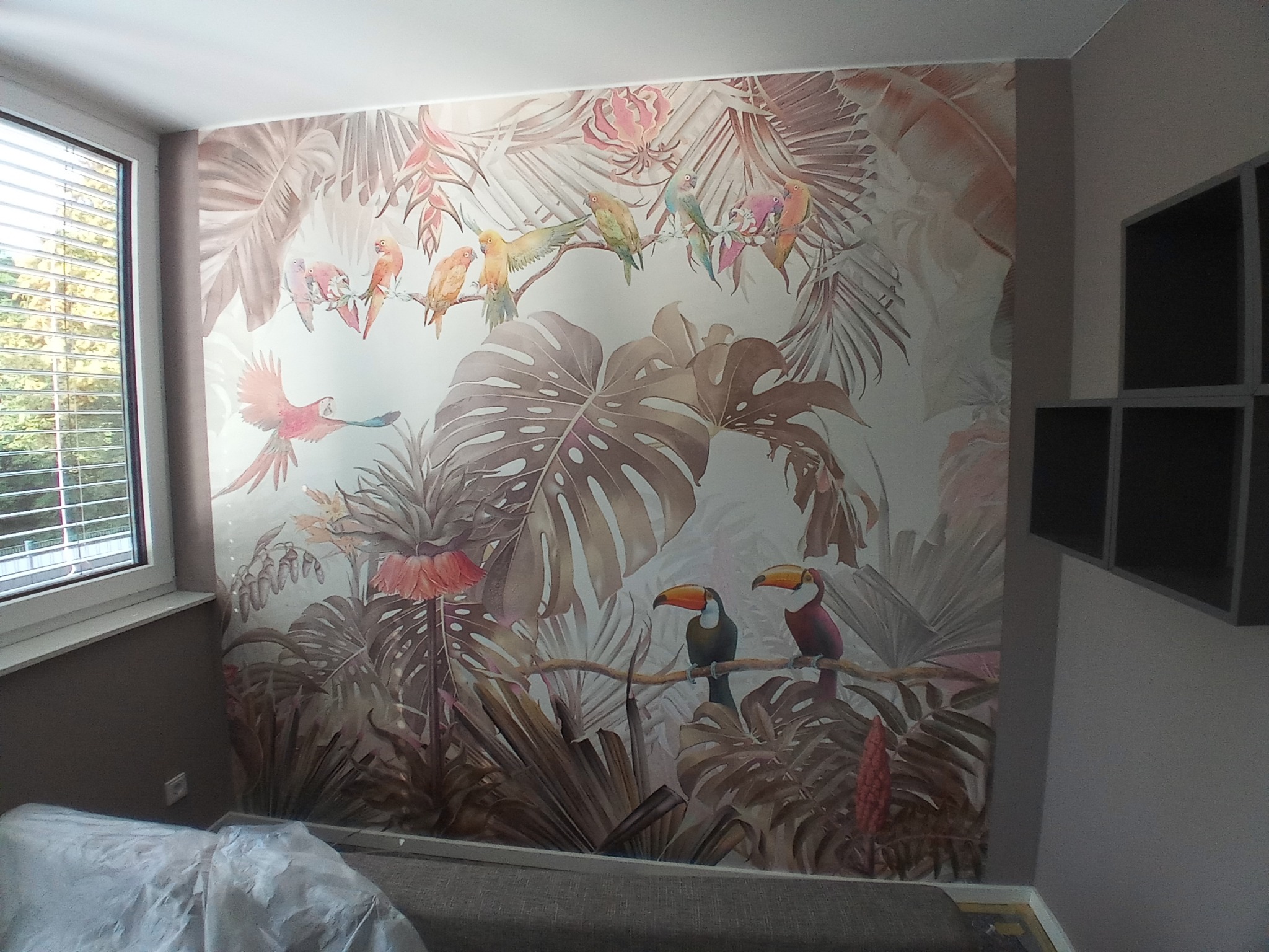Ściana z fototapetą przedstawiającą tropikalny motyw z tukanami i papugami, widoczna częściowo zasłonięta folią podłoga i fragment regału na ścianie obok okna.