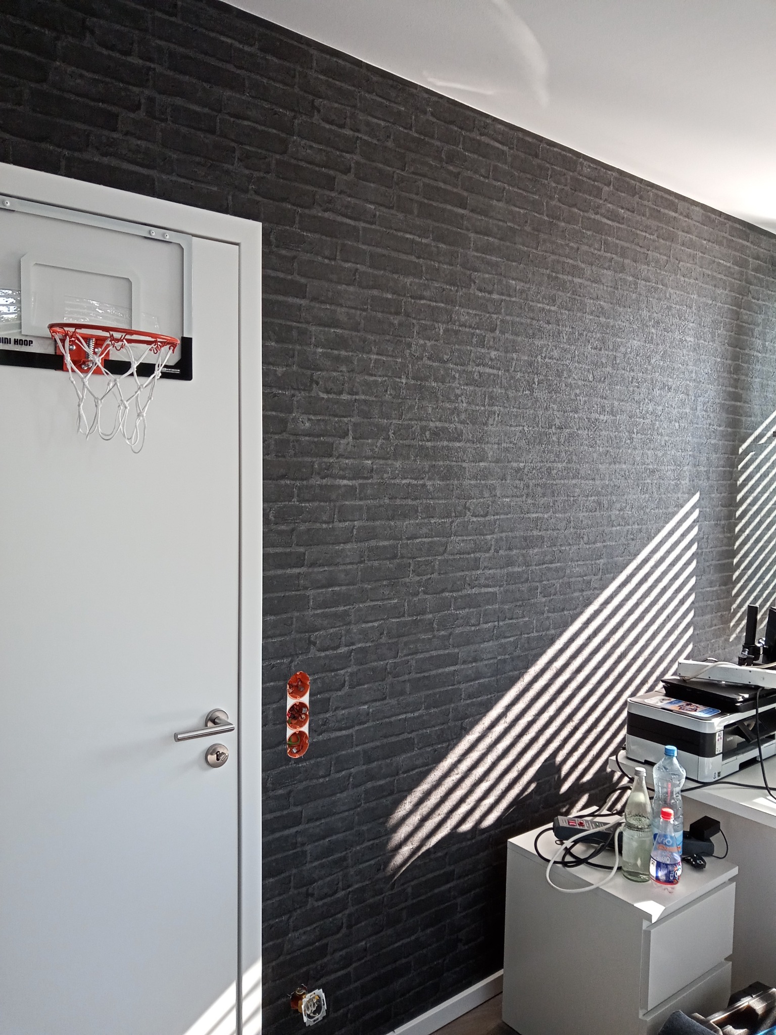 Pokój z ciemną ścianą imitującą cegłę, białe drzwi z mini koszem do koszykówki i biurko z drukarką, oświetlone promieniami słońca wpadającymi przez żaluzje.