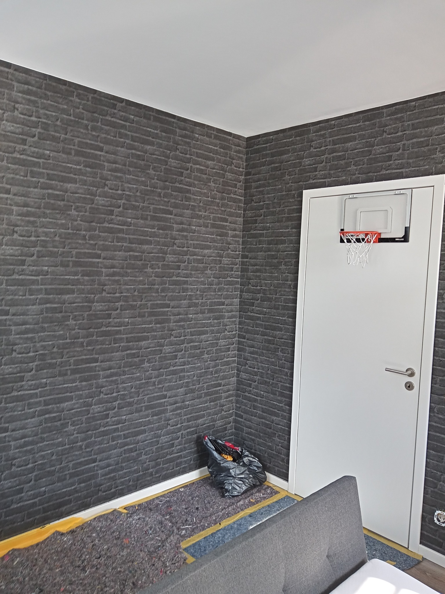 Ściana wykończona szarą cegłą dekoracyjną, podłoga w trakcie przygotowania do położenia paneli, białe drzwi z mini koszem do koszykówki.