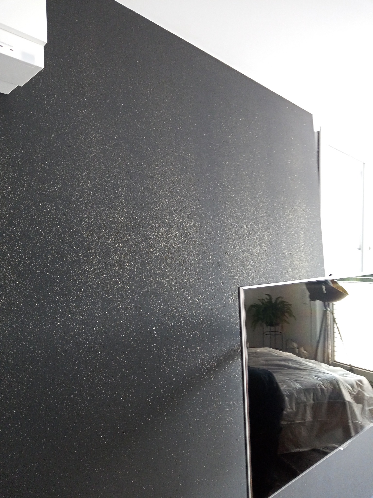 Ściana pomalowana na ciemny kolor z delikatnym, złotym połyskiem widoczna w perspektywie, odbicie sypialni w lustrze obok.