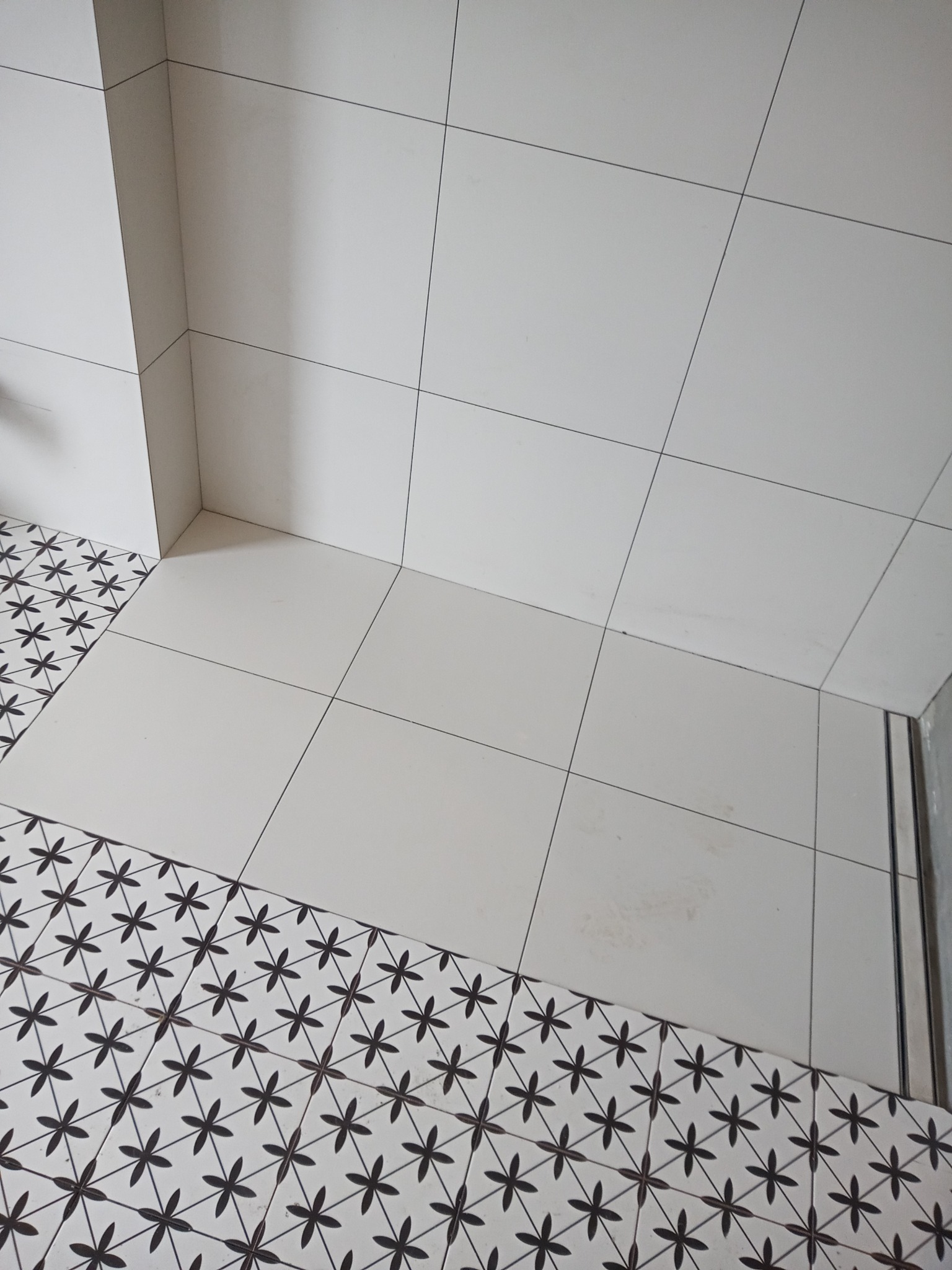 Wykończona łazienka z białymi kwadratowymi płytkami ściennymi i podłogą z dekoracyjnymi, czarno-białymi płytkami w kwiatowy wzór, widoczne łączenie płytek pod kątem.