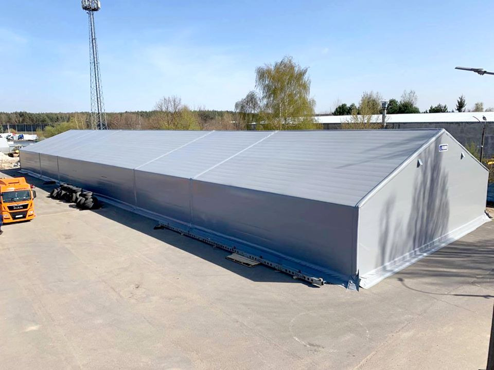 Duża, szara hala magazynowa z dachem dwuspadowym, widoczna z zewnątrz na betonowym placu, z pomarańczową ciężarówką MAN w lewym dolnym rogu i stosem opon obok. W tle drzewa i słup wysokiego napięcia.