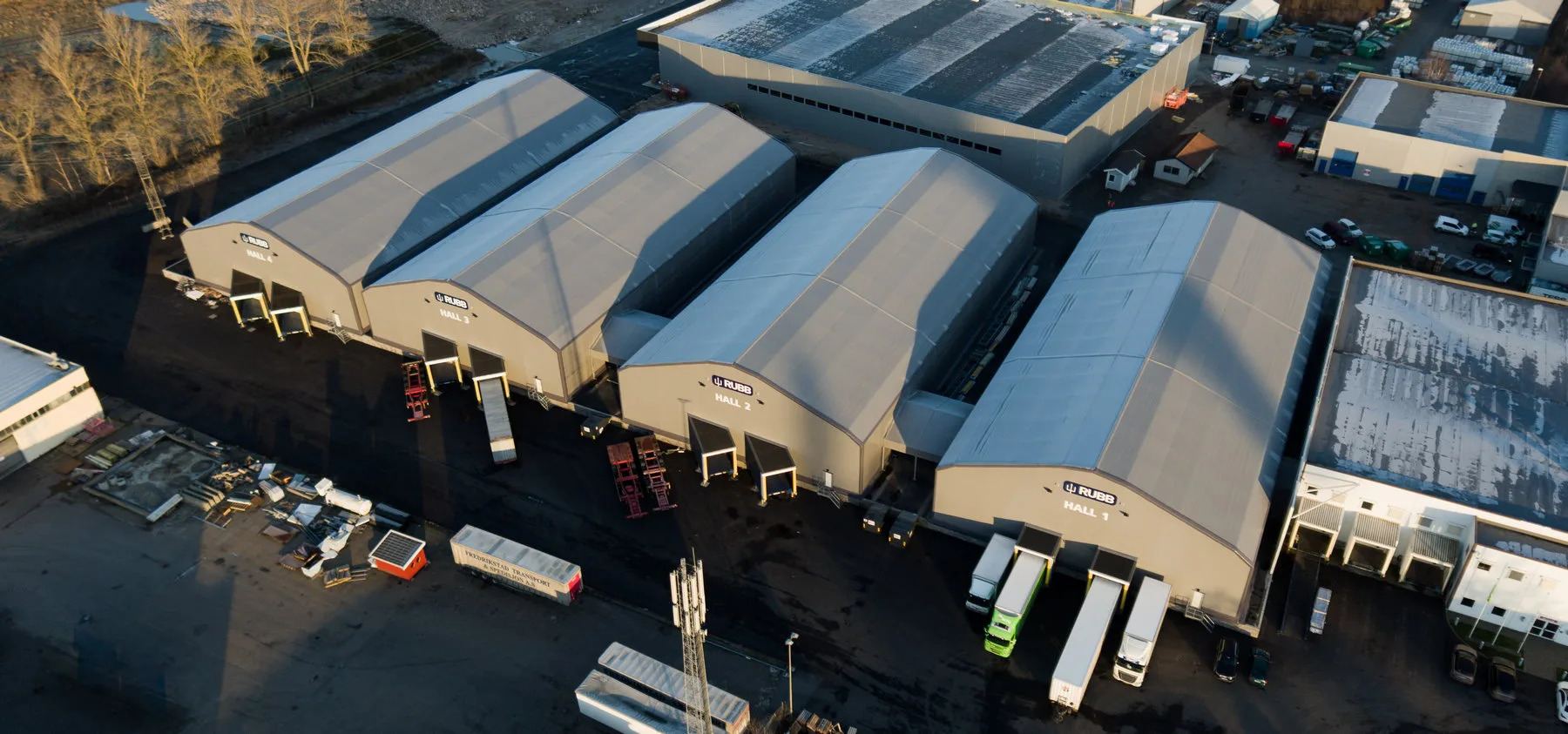 Widok z lotu ptaka na cztery hale magazynowe z szarej plandeki z logo RUBB, ustawione szeregowo, z rampami załadunkowymi i zaparkowanymi ciężarówkami, w tle zabudowania przemysłowe i drzewa.