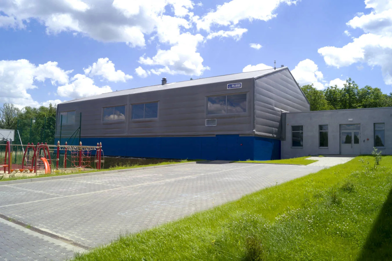 Hala sportowa z szarej membrany z niebieską podmurówką, przylegająca do szarego budynku, widoczny plac zabaw i parking