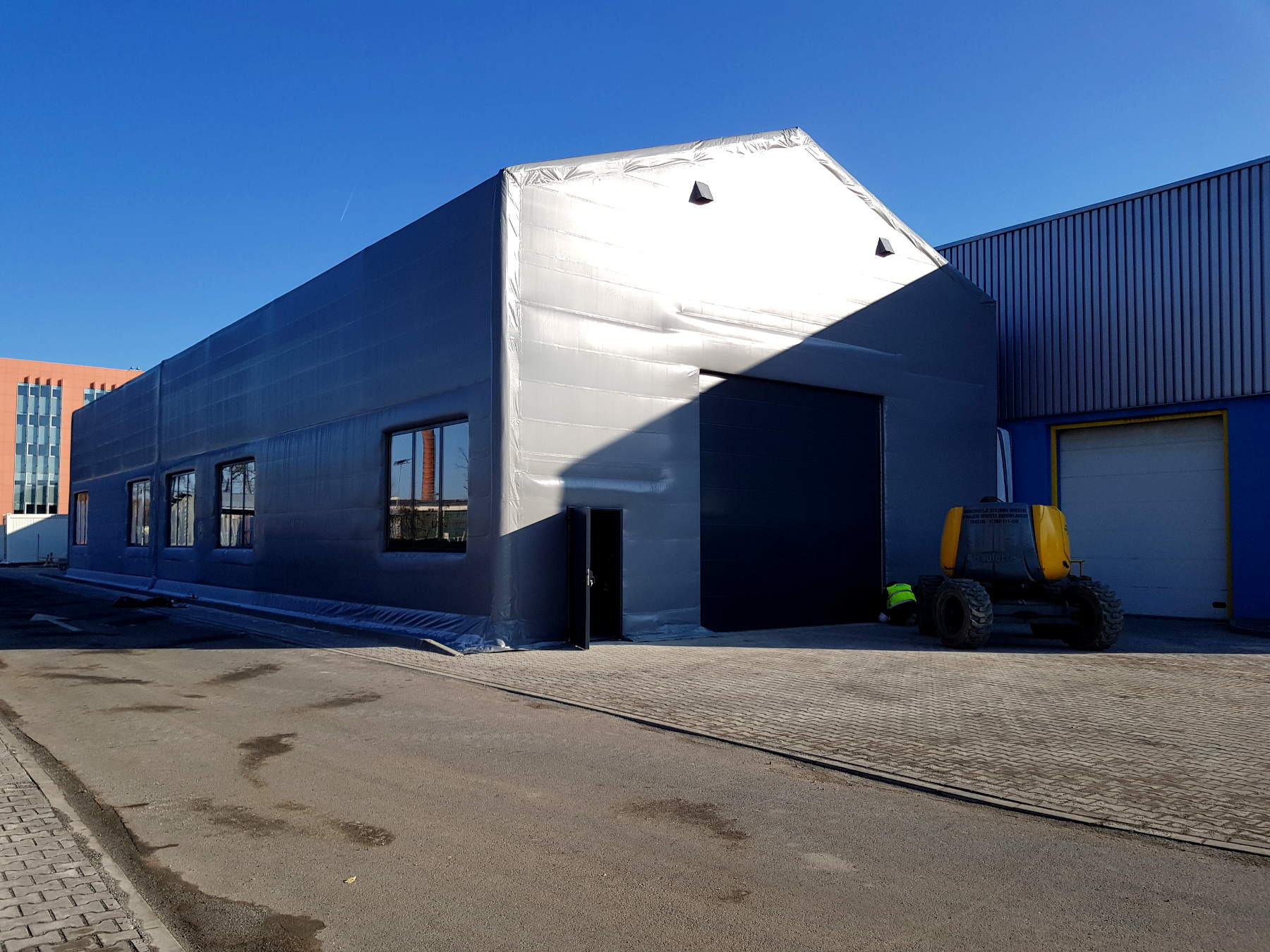 Zewnętrzny widok hali magazynowej o konstrukcji stalowej, pokrytej szarym materiałem plandekowym, z widocznymi oknami, drzwiami i bramą wjazdową. Obok stoi żółta maszyna budowlana, a w tle widać...