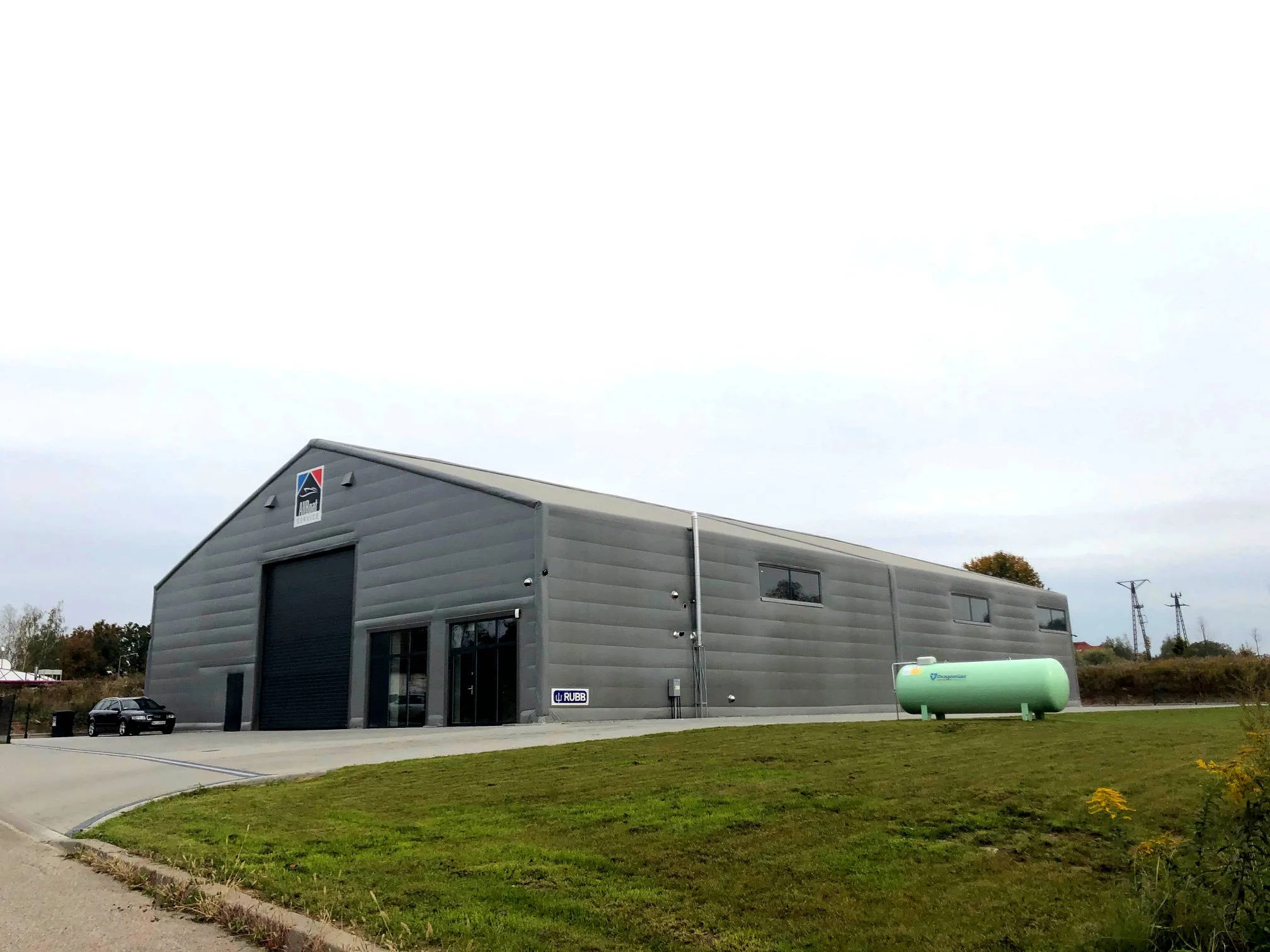 Szara hala magazynowa z logo firmy AllBoat i zielonym zbiornikiem DragonGaz na trawniku przed budynkiem, widok z zewnątrz w pochmurny dzień.