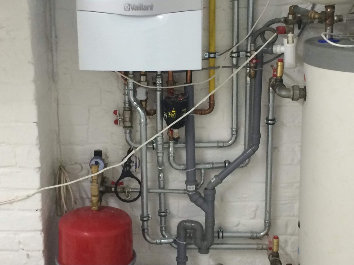 Kocioł gazowy Vaillant z widocznymi rurami instalacji grzewczej na tle ściany z białej cegły, z czerwonym zbiornikiem przeponowym na pierwszym planie.