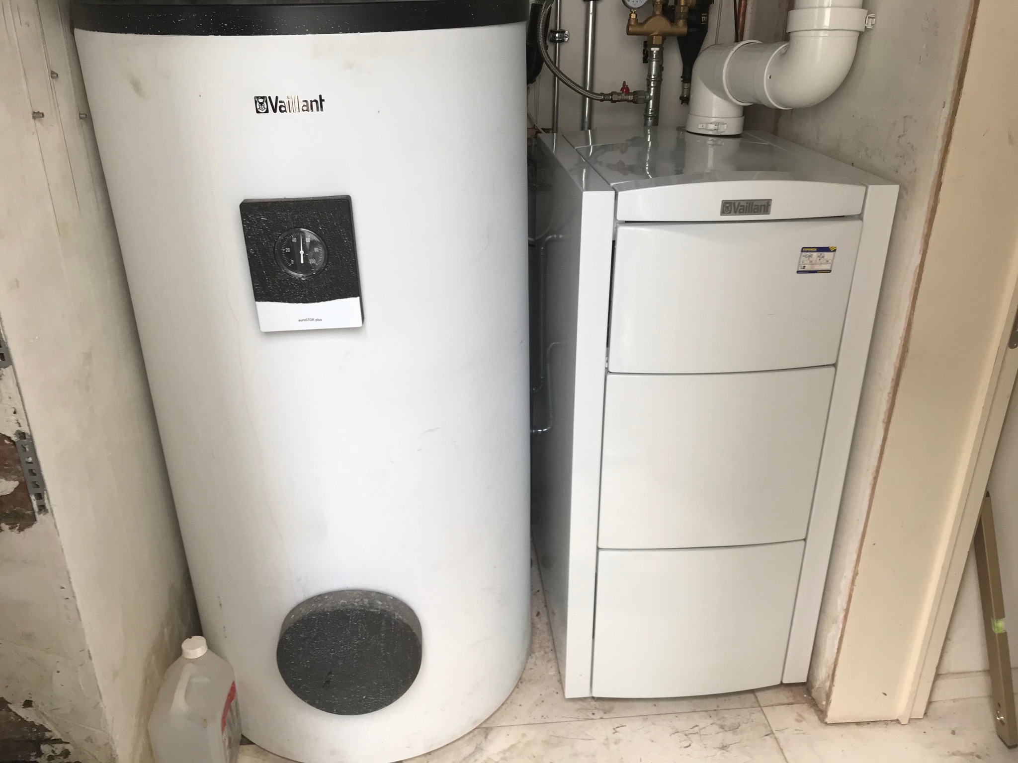 Biały, kondensacyjny kocioł gazowy Vaillant z zasobnikiem ciepłej wody, widoczne rury i zawory, zamontowane w pomieszczeniu ze ścianami w trakcie remontu.