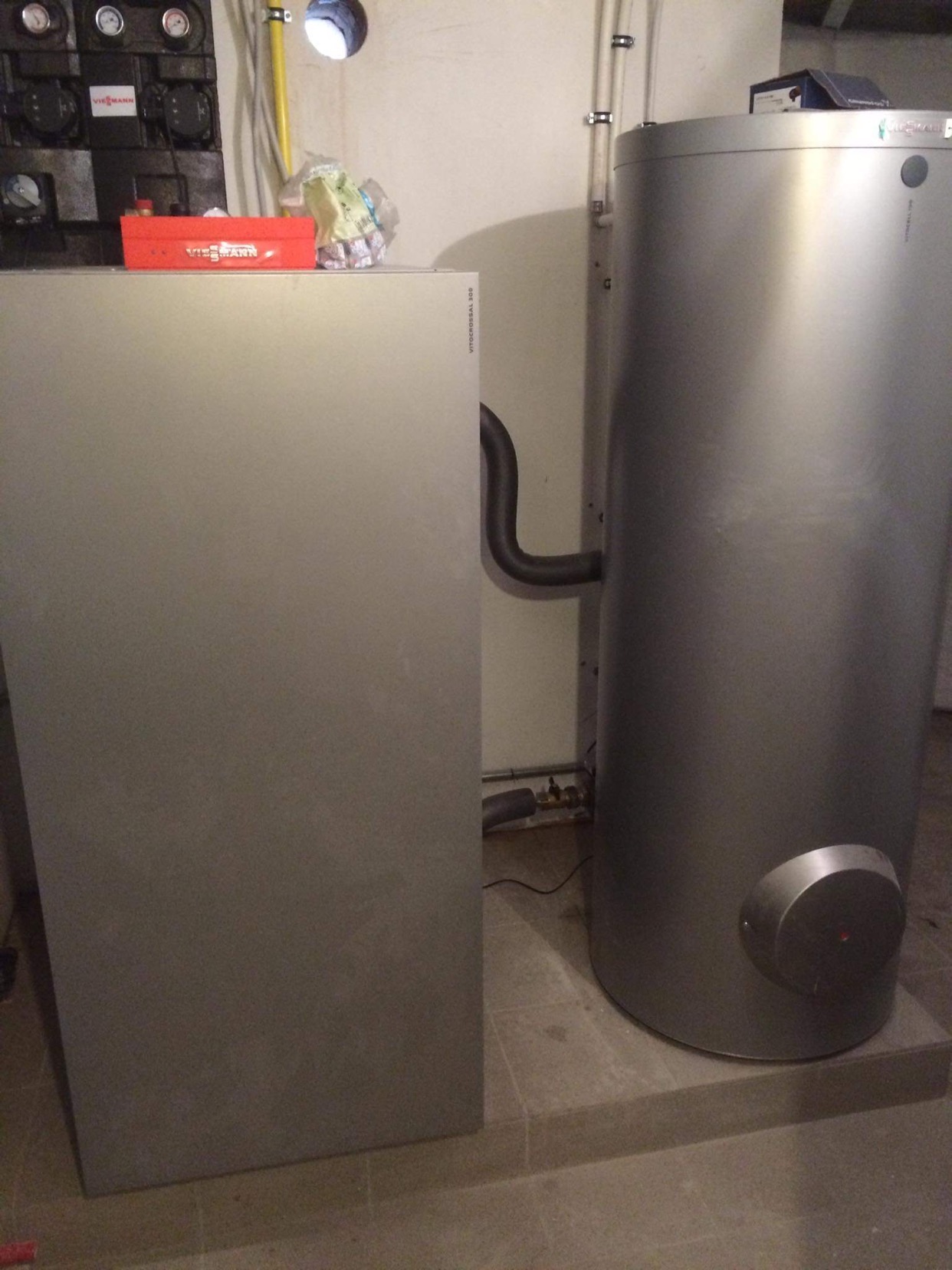 Szara kotłownia z dwoma zbiornikami, z których jeden jest pionowy i cylindryczny, a drugi płaski i prostokątny, z czerwonym pudełkiem narzędzi Viessmann na górze płaskiego zbiornika.