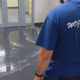 Pracownik firmy sprzątającej Woj&Mar w niebieskiej koszulce czyści podłogę z płytek w biurze, widoczne ślady piany po myciu.