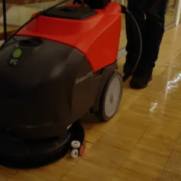 Automat szorująco-zbierający IPC Gansow CT15 na drewnianej podłodze po umyciu, widoczne ślady wilgoci, w tle schody i osoba obsługująca maszynę.