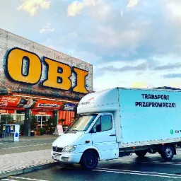 Biały samochód dostawczy firmy przeprowadzkowej zaparkowany przed sklepem OBI z widocznym logo firmy na boku pojazdu i numerem telefonu.