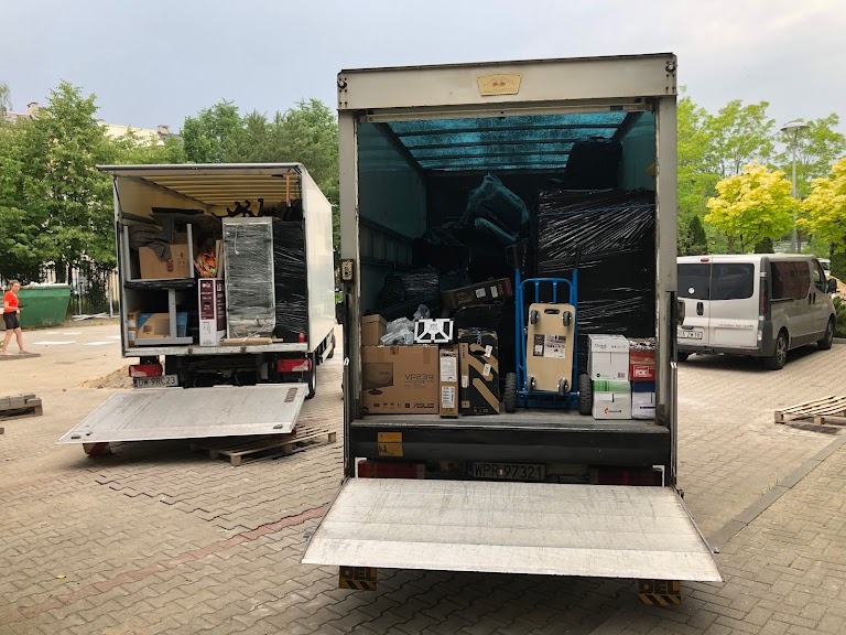 Dwa samochody dostawcze z otwartymi rampami załadowane meblami i sprzętami, przygotowane do transportu podczas przeprowadzki.