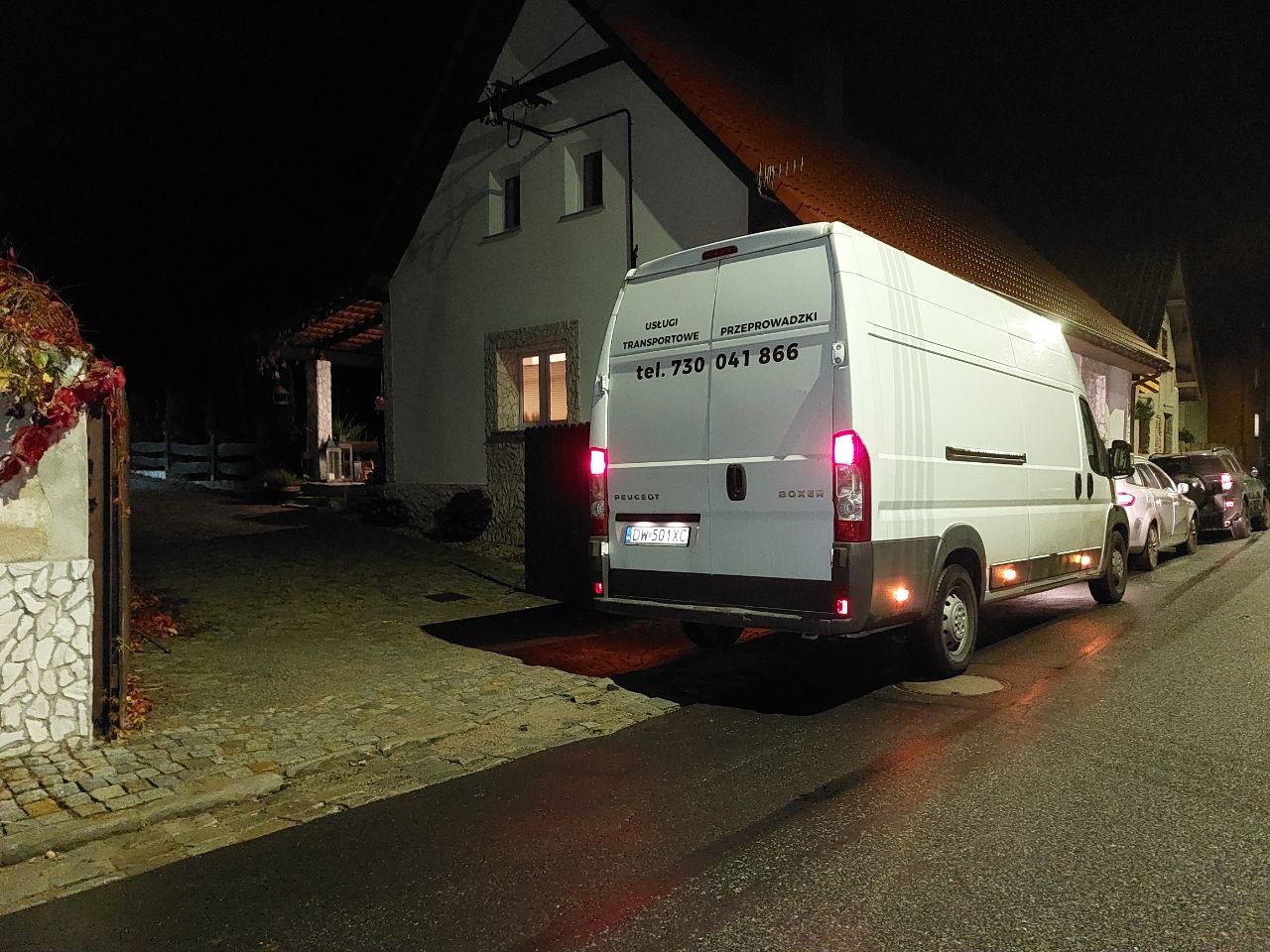Biały samochód dostawczy Peugeot Boxer z napisem 'Usługi Transportowe Przeprowadzki' i numerem telefonu zaparkowany nocą przed domem.