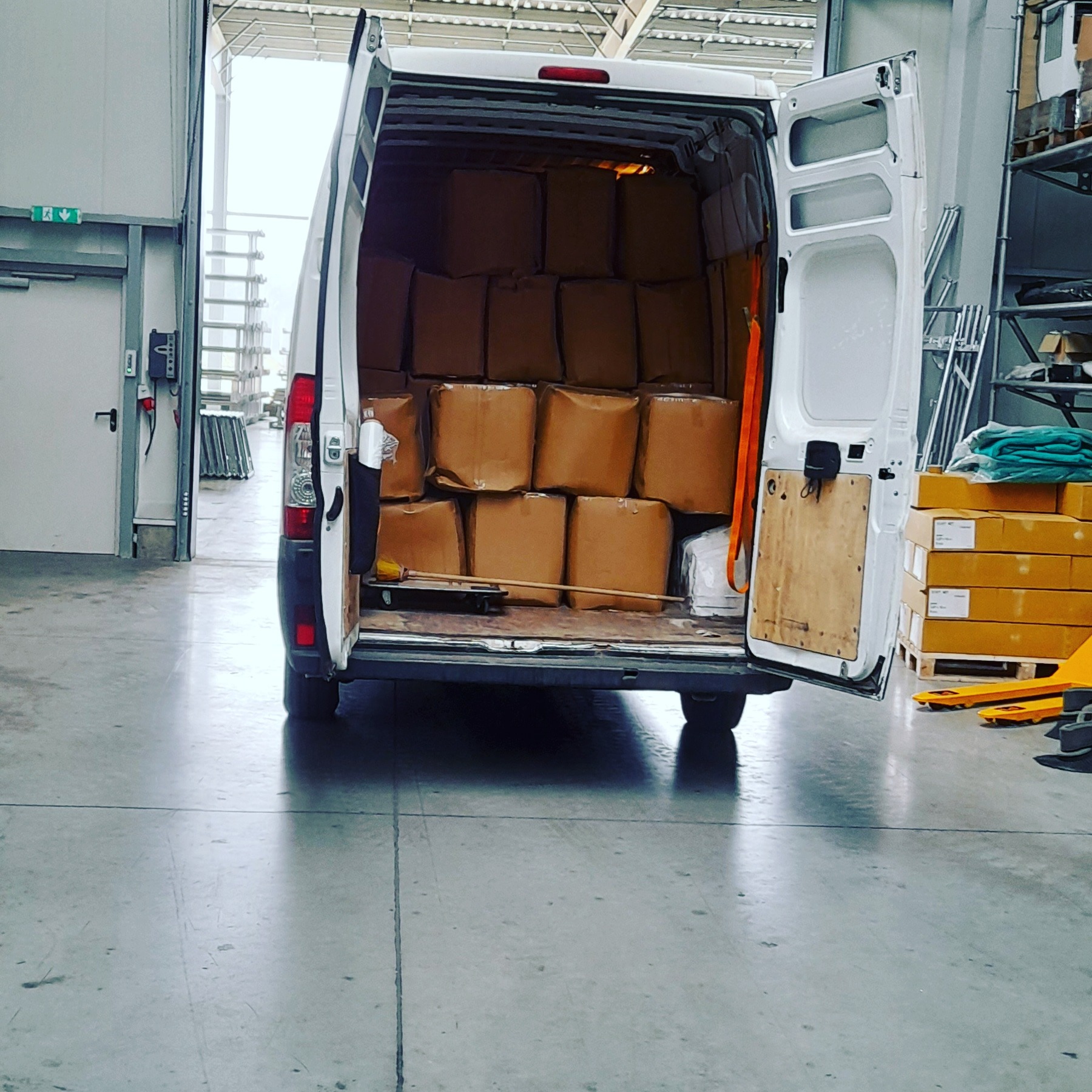 Wnętrze białego furgonetki wypełnione kartonami, widok z tyłu, z otwartymi drzwiami, w tle magazyn z regałami i paletami.