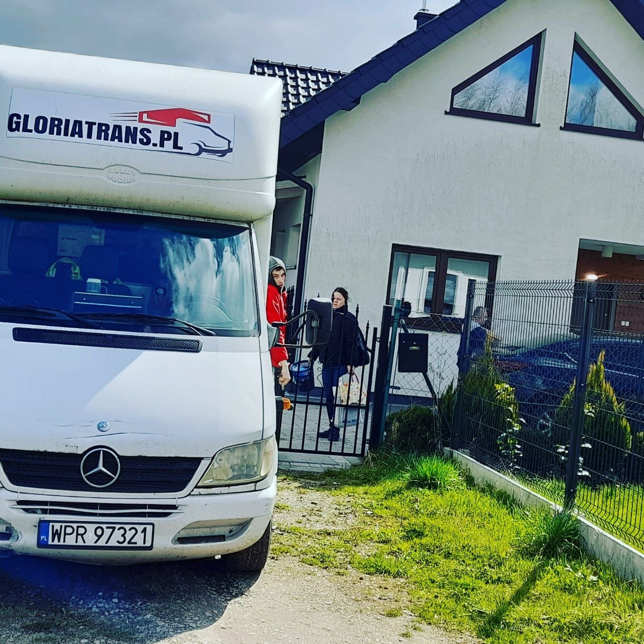 Biały samochód dostawczy firmy GLORIATRANS.PL zaparkowany przed domem z trójkątnymi oknami, osoby wchodzą przez furtkę.