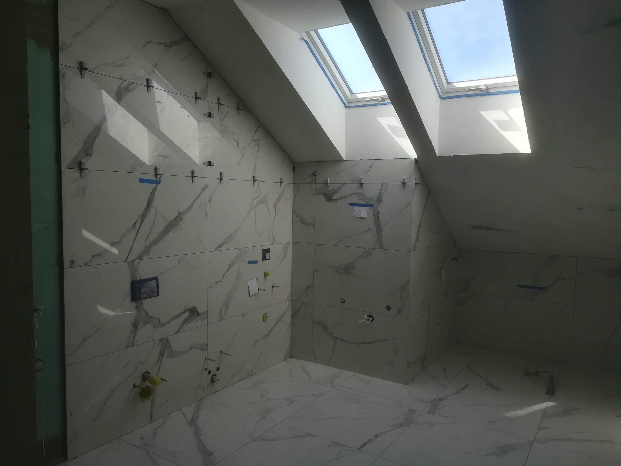 Wnętrze łazienki na poddaszu w trakcie układania płytek imitujących marmur, widoczne krzyżyki dystansowe, okno dachowe i przygotowane instalacje wodno-kanalizacyjne.