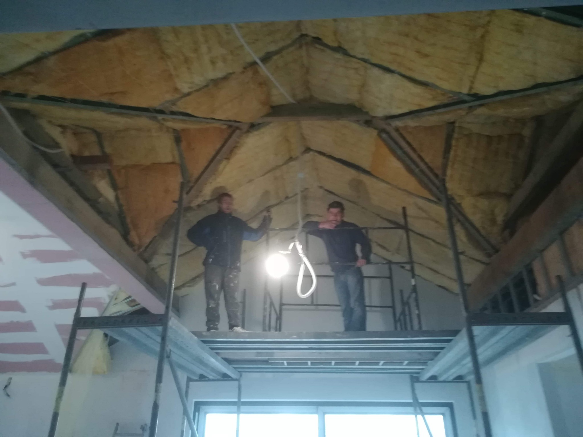 Dwaj mężczyźni na rusztowaniu montują izolację z wełny mineralnej na poddaszu, widoczne drewniane krokwie i elementy konstrukcyjne dachu, tymczasowe oświetlenie budowlane.