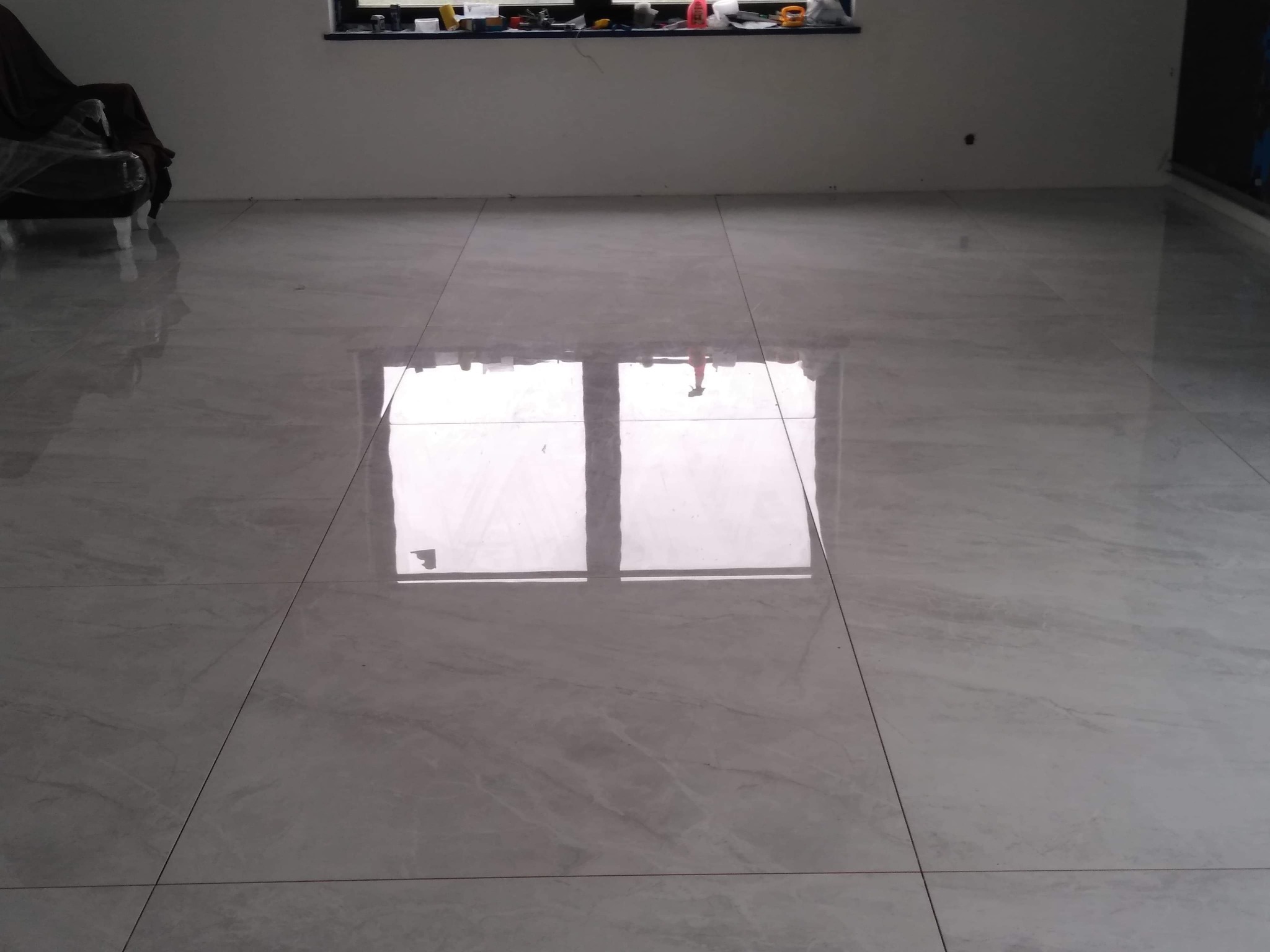 Błyszcząca, jasnoszara podłoga z płytek ceramicznych, widoczne fugi, odbicie okna, fotel przy ścianie, narzędzia na parapecie okiennym.