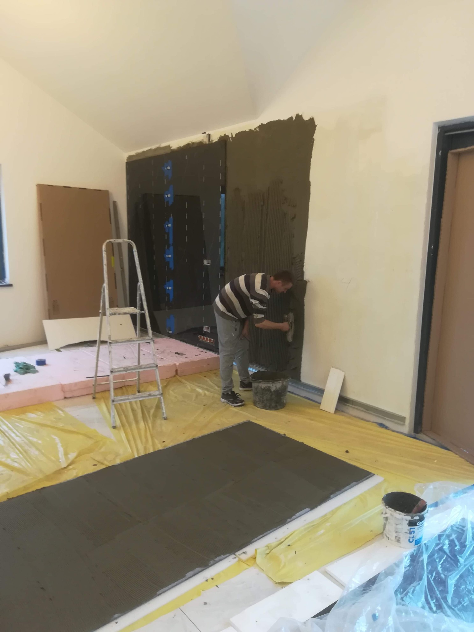 Pracownik w Grodzisku Mazowieckim nakłada klej na ścianę w pomieszczeniu przygotowywanym do położenia płytek, widoczne narzędzia, wiadro z klejem, fragmenty płyt oraz zabezpieczona folią podłoga.