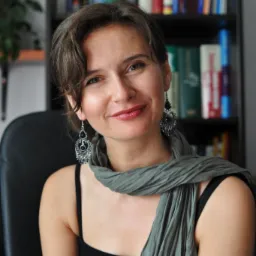 Portret uśmiechniętej kobiety z kolczykami i szalikiem na tle regału z książkami.