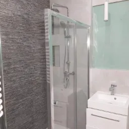 Nowoczesna łazienka po remoncie: szara ściana z teksturą, kabina prysznicowa z chromowanymi elementami, biała szafka z umywalką i pionowa lampa wisząca.