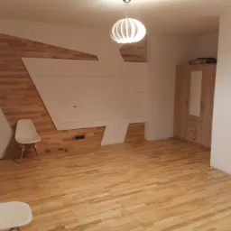 Jasne panele podłogowe ułożone w nowym pokoju z drewnianą ścianą z geometrycznym wzorem, minimalistyczne krzesła i nowoczesna lampa wisząca.