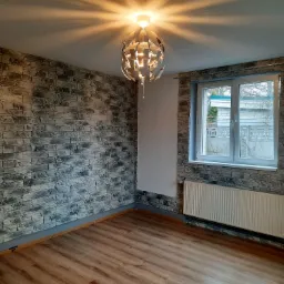 Wnętrze pokoju po remoncie: ściana pokryta tapetą imitującą kamień, drewnopodobne panele podłogowe, biały grzejnik pod oknem i designerska lampa wisząca.