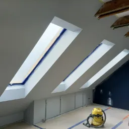 Poddasze z trzema oknami dachowymi po świeżym zagruntowaniu ścian, widoczny pracownik na drabinie i żółty odkurzacz przemysłowy.