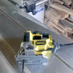 Żółto-czarna wyrzynarka Stanley Fatmax leży na metalowym stole warsztatowym, w tle stos drewnianych odpadów i narzędzia.