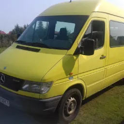 Żółty bus Mercedes-Benz 312D, zaparkowany na trawie, z widoczną tablicą rejestracyjną i logo firmy na boku.