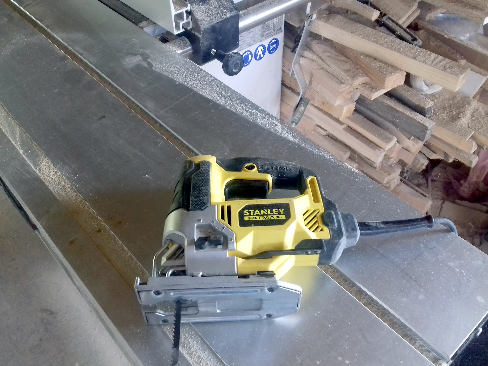 Żółto-czarna wyrzynarka Stanley Fatmax leży na metalowym stole warsztatowym, w tle stos drewnianych odpadów i narzędzia.