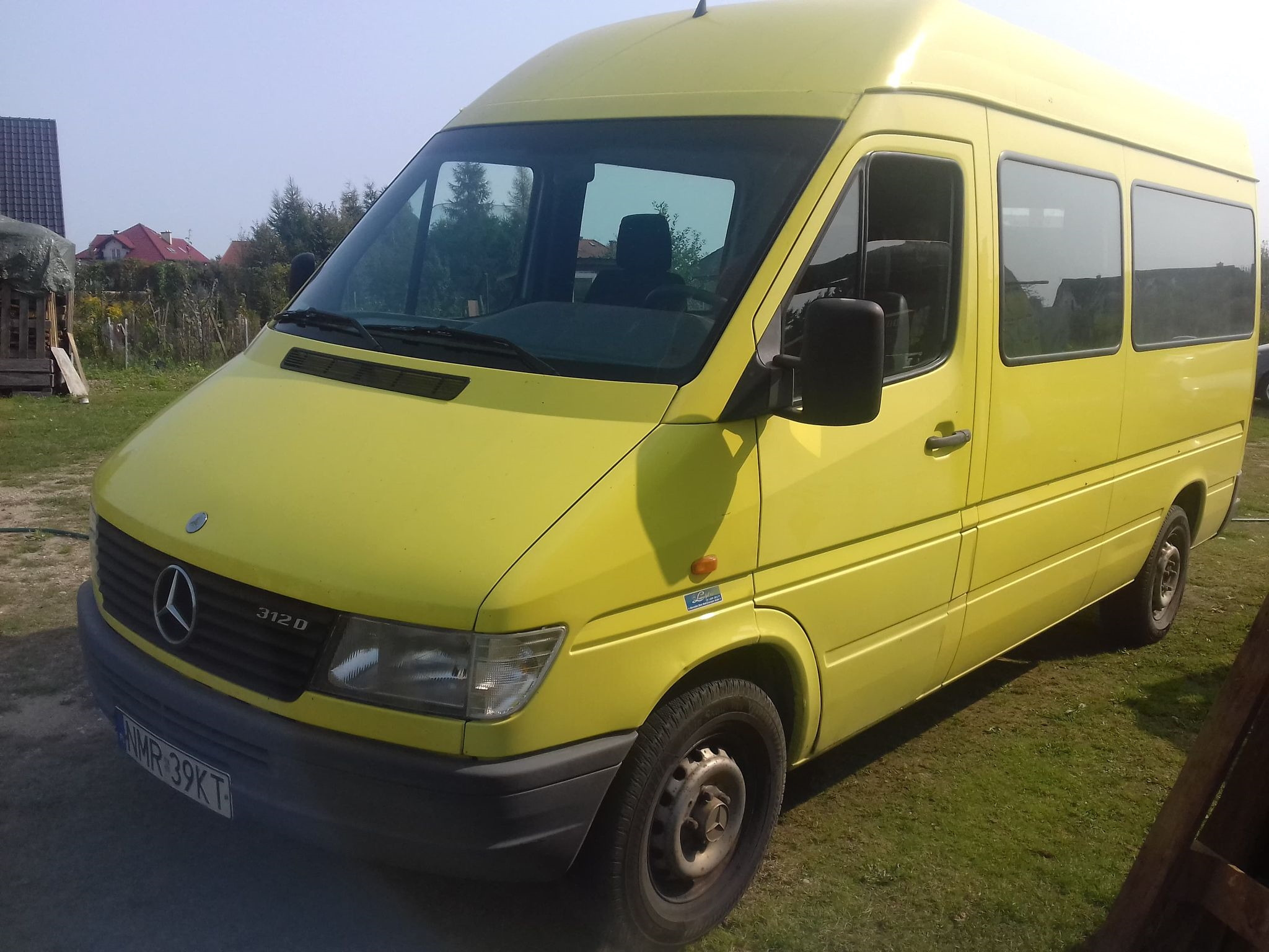 Żółty bus Mercedes-Benz 312D, zaparkowany na trawie, z widoczną tablicą rejestracyjną i logo firmy na boku.