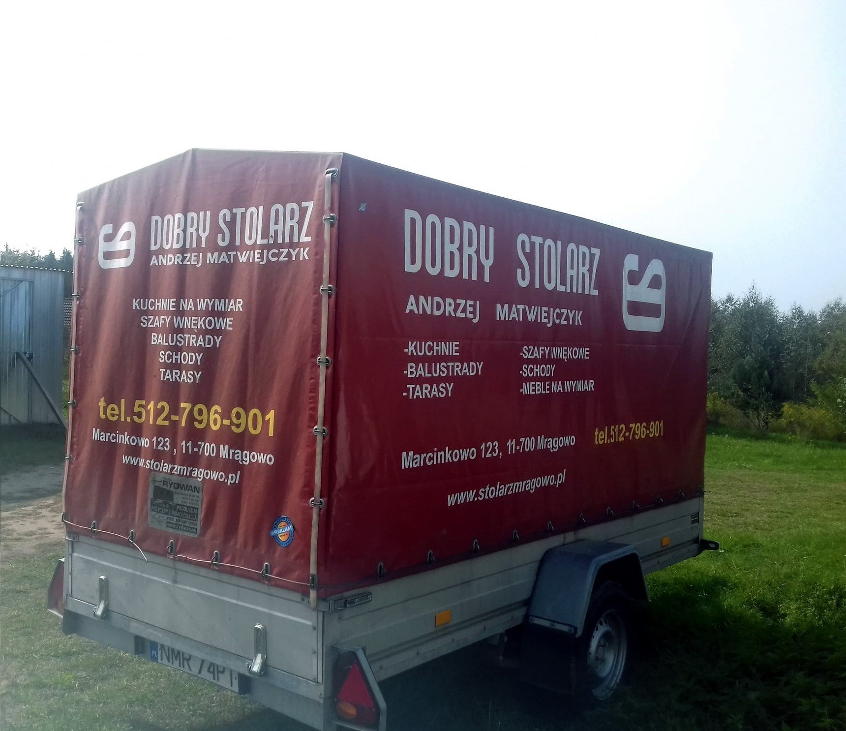 Przyczepka samochodowa z reklamą usług stolarskich: kuchnie na wymiar, szafy wnękowe, balustrady, schody, tarasy, z adresem i numerem telefonu firmy Dobry Stolarz Andrzej Matwiejczyk z Mrągowa.