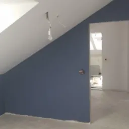 Pomalowane na granatowo poddasze ze skośnym sufitem i widoczną żarówką bez klosza, surowy beton na podłodze, widok na kolejne pomieszczenie przez otwór drzwiowy bez drzwi.