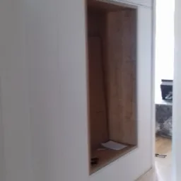 Wnęka w zabudowie meblowej z drewnianym wnętrzem, widoczne elementy montażowe i kartka papieru w środku, całość w jasnej kolorystyce.