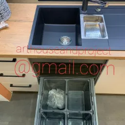 Czarny, kompozytowy zlew kuchenny z ociekaczem i baterią na blacie z drewnianym dekorem; wysuwana szuflada na kosze do segregacji odpadów z jednym koszem wypełnionym workiem foliowym.