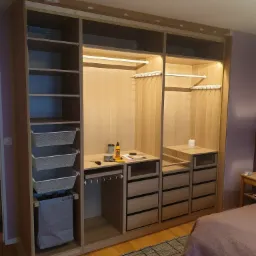 Wnętrze szafy wnękowej w trakcie montażu, widoczne p&oacute;łki, kosze, drążki, szuflady, oświetlenie LED, narzędzia i klej na blacie roboczym, zabezpieczenia taśmą malarską na g&oacute;rze.