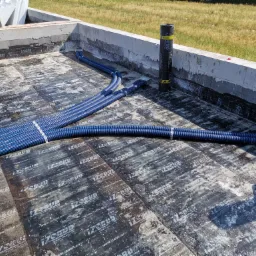 Niebieskie rury karbowane, prawdopodobnie wentylacyjne, ułożone na zaizolowanej powierzchni fundamentu budynku w trakcie budowy, z widocznymi rolkami materiału izolacyjnego i fragmentem muru.